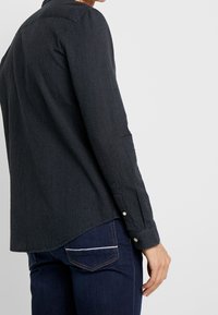 Camicia nera con bottoni, caratterizzata da un piccolo motivo a pois bianchi, con maniche lunghe e orlo curvo. Il materiale sembra liscio e strutturato.