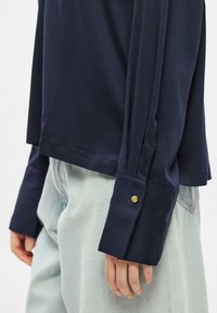 Μακρυμάνικο πουκάμισο σε ναυ�τικό μπλε από απαλό ύφασμα, με μανσέτα που κουμπώνει και διακόσμηση σε χρυσό, συνδυασμένο με ανοιχτόχρωμο τζιν παντελόνι.