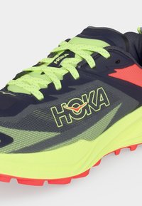 Ναυτικό μπλε και νέον κίτρινο αθλητικό παπούτσι HOKA με φωτεινά κίτρινα κορδόνια, νέον κίτρινη μεσαία σόλα και κόκκινο πέλμα εξωτερικής σόλας.