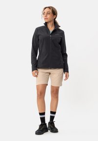 Zwarte softshelljack met hoge kraag, ritssluiting en logo. Gecombineerd met beige shorts, zwarte schoenen en zwart-witte gestreepte sokken.