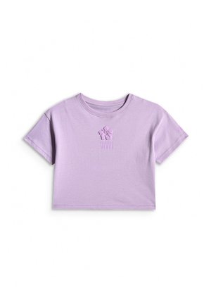 Dziewczyna stojąca, ubrana w fioletowy t-shirt z krótkim rękawem, beżowe szorty, białe skarpetki i białe trampki na szarym tle.