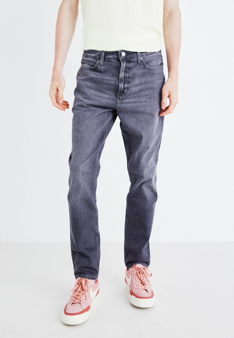 Moške sive denim hlače s ravnim krojem, opremljene s petimi žepi, gumbom za zapiranje in rahlo zbledelim materialom. V kombinaciji z roza športnimi copati.