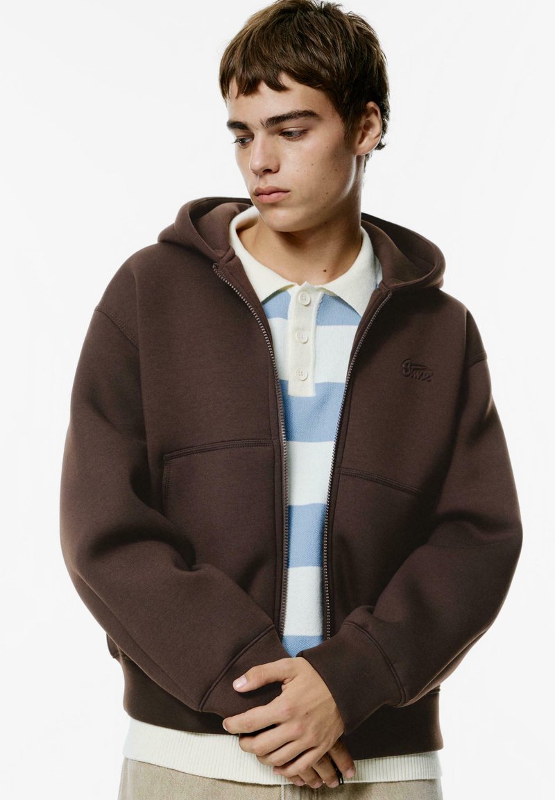 Bruine zip-up hoodie met een zachte textuur, voorzakken en een capuchon met trekkoord, gelaagd over een lichtblauw gestreept poloshirt.