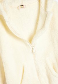 Veste à capuche zippée en polaire couleur crème, présentant une texture douce, de grandes poches avant et une capuche confortable, conçue pour les enfants.