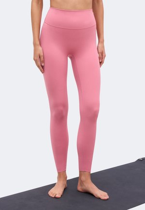 Osoba stoi boso na czarnej macie do jogi, ubrana w różowe legginsy z wysokim stanem o pełnej długości na jednolitym tle.