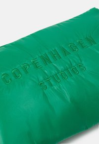 Coussin vert rembourré avec le texte en relief "COPENHAGEN STUDIOS", affichant une texture brillante et une surface matelassée.