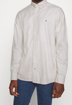 Hombre con camisa blanca de manga larga abotonada con un pequeño logo de Tommy Hilfiger en el pecho y vaqueros azules.