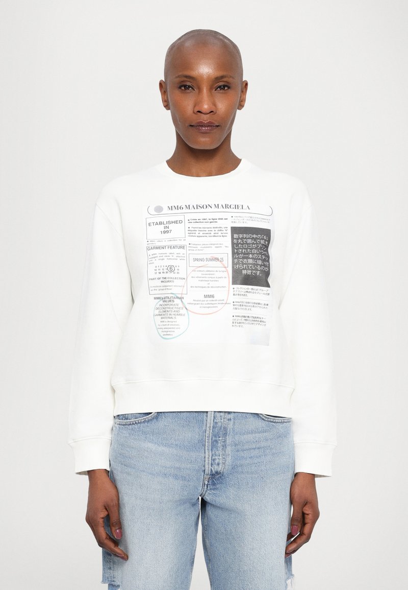 White sweatshirt with printed labels and text in various languages. Crew neck, long sleeves, ribbed cuffs, and relaxed fit. Worn with light blue jeans.