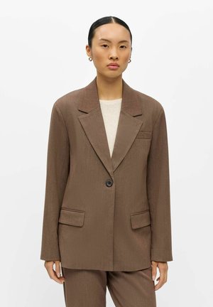 Vrouw met naar achter geslagen haar, die een bruine oversized blazer en bijpassende broek draagt over een lichtgekleurde top, staand tegen een effen achtergrond.