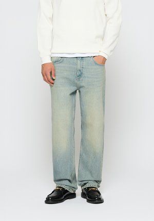 Άνδρας που φοράει ελαφρώς μπλε τζιν με ίσια γραμμή, λευκό μακρυμάνικο μπλουζάκι και μαύρα μοκασίνια με λεπτομέρεια χρυσής αλυσίδας, στέκεται με το ένα χέρι στην τσέπη.