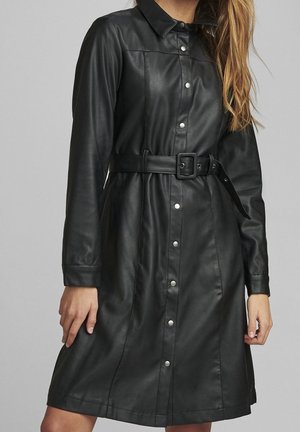 Femme portant une robe noire ceinturée en similicuir avec manches longues et boutons-pression sur le devant, contre un fond uni.