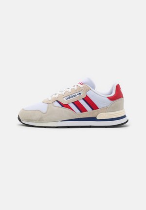 Baskets blanches Adidas avec des bandes rouges, des accents en suède beige, un détail bleu sur la semelle intermédiaire et des lacets blancs, vues de côté sur un fond gris.
