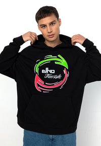 Schwarzer Hoodie mit einem großen kreisförmigen Grafikdesign, das rote, grüne und weiße Wirbel zeigt. Vorderseitige Kängurutasche, lockere Passform und verstellbare Kapuze.