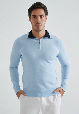 Pull bleu clair à manches longues avec un col zippé. Le col est bleu marine avec un accent contrastant et des poignets et un ourlet côtelés.
