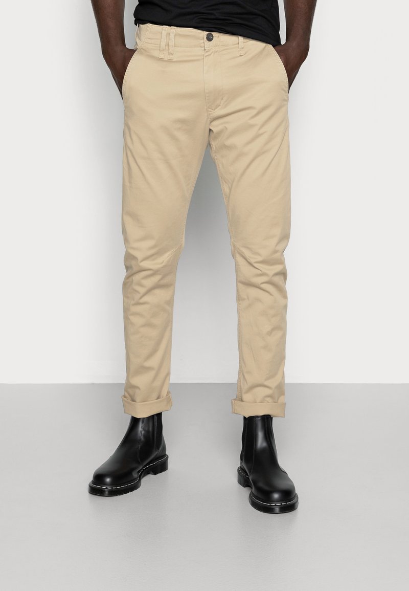Homme portant un chino beige retroussé et des bottines en cuir noir, mains dans les poches, debout devant un fond clair uni.