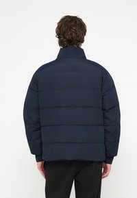 Tmavomodrá prešívaná bunda s vysokým golierom, s horizontálnym prešívaním, elastickými manžetami a voľným strihom. Hladká, textúrovaná látka.