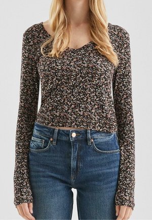 Femme portant un haut noir à manches longues à motif floral et un jean bleu taille haute, debout devant un fond clair uni.
