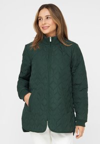 Grüne gesteppte Jacke mit hohem Kragen, Reißverschluss, gerippten Bündchen und zwei seitlichen Taschen. Das Muster zeigt rautenförmige Steppnähte.