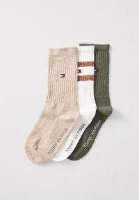 Tre par med ribbestrikkede sokker: lys beige, hvit med brune striper, og mørkegrønn. Hver av dem har en logo på ankelen og strukturert stoff.