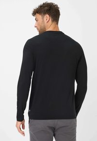 Schwarzes Langarmshirt aus weichem Stoff mit Rundhalsausschnitt und einem kleinen Markenetikett am Saum, getragen mit grauen Hosen.