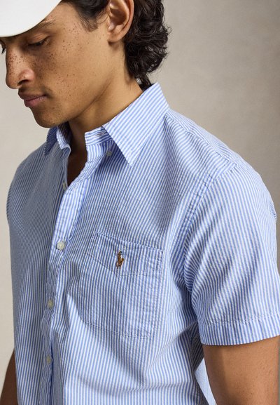 Homme aux cheveux bouclés portant une casquette blanche et une chemise à manches courtes rayée bleu et blanc avec une poche poitrine et un logo brodé.