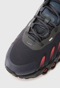 Tênis Nike com parte superior de malha preta e detalhes cinzas, realces em vermelho e bico arredondado. Inclui uma sola acolchoada com sulcos pronunciados.