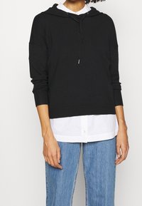 Sweatshirt noir à capuche avec des poignets et un ourlet en côte, superposé à une chemise blanche à boutons. Le tissu semble doux et légèrement texturé.