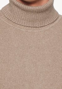 Beige stickad turtlenecktröja med en ribbad krage som visas på en persons övre bröst och nacke.