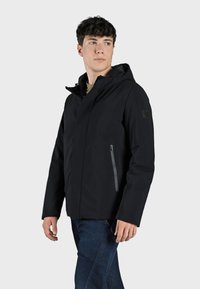 Giacca nera impermeabile con tasca con zip, cappuccio regolabile e design elegante, abbinata a jeans blu scuro.