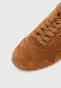 Καφέ σουέντ αθλητικό παπούτσι με στρογγυλερή μύτη, υφασ μένο επάνω μέρος, δέσιμο με κορδόνια και λαστιχένια σόλα. Διαθέτει διακριτικές ραφές και λείες λεπτομέρειες.