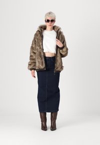 Ψεύτικο γούνινο μπουφάν σε καφέ αποχρώσεις, κομμένο λευκό τοπ, σκούρα τζιν φούστα και καφέ μποτάκια με τακούνι. Το μοντέλο φορά γυαλιά ηλίου και ποζάρει όρθιο.