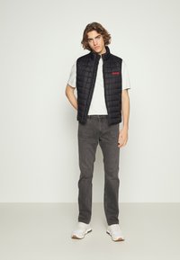 Gilet trapuntato nero con colletto alto e logo rosso "HUGO", indossato sopra una maglietta chiara, abbinato a jeans scuri e sneakers bianche.