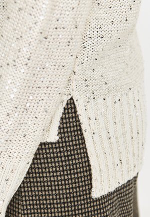 Sweter dzianinowy w kremowym kolorze z srebrnymi cekinami, z prążkowanymi brzegami i bocznymi rozcięciami, nałożony na brązowy materiał w kratkę.