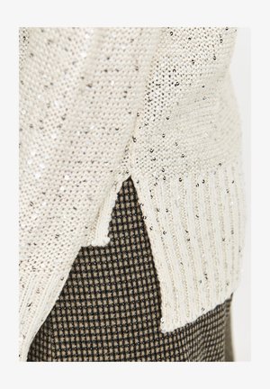 Sweter dzianinowy w kremowym kolorze z srebrnymi cekinami, z prążkowanymi brzegami i bocznymi rozcięciami, nałożony na brązowy materiał w kratkę.