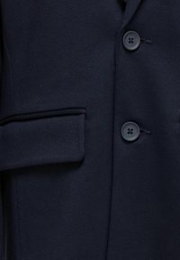 Blazer bleu marine foncé en tissu lisse, comportant deux boutons et une poche poitrine simple avec une finition soignée. La texture semble douce et structurée.