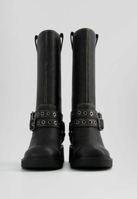 Botas altas hasta la rodilla de cuero negro con ojales metálicos redondos y detalles de hebilla. Presentan una suela gruesa y un diseño recto y estructurado.