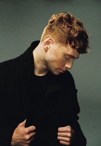 Porträtt av en person med kort, lockigt rött hår, iklädd en svart kappa, med händerna ihop och huvudet lätt böjt, mot en neutral bakgrund.