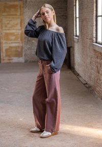 Grijze, gestructureerde top met schoudervullingen, gecombineerd met roze wijde broek en witte sneakers, tegen een rustieke binnenachtergrond.