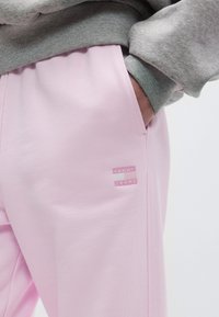 Άτομο που φοράει ανοιχτό ροζ παντελόνι φόρμας Tommy Jeans με το χέρι στην πλαϊνή τσέπη, σε συνδυασμό με μανίκι γκρι φούτερ ορατό.