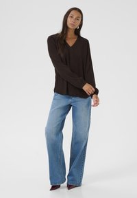 Blusa nera a maniche lunghe con scollatura a V, abbinata a jeans a gamba larga blu chiaro. Modella in scarpe con punta bordeaux. Tessuto dalla texture liscia.