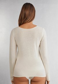 Pull à manches longues de couleur crème avec un col rond et une texture douce. Il comporte un ourlet asymétrique et des détails en côtes sur les bords.
