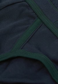 Marinblått tyg som blandas med gröna elastiska detaljer; slät textur med sydda kanter och en subtil lyster, vilket betonar en modern design.