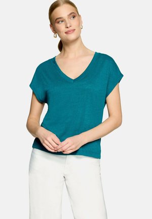 Femme aux cheveux blonds en queue de cheval portant des boucles d'oreilles créoles dorées, un haut bleu sarcelle à col en V manches courtes, et un pantalon blanc, debout les mains jointes.