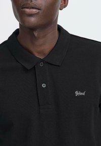 Nahaufnahme einer Person, die ein schwarzes Polohemd mit zwei Knöpfen und einem weißen, auf der Brust gestickten "Blend"-Logo trägt.