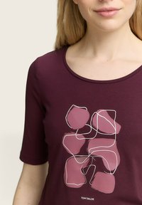 Burgunderfarbenes T-Shirt mit weitem Ausschnitt und ärmellangen Ärmeln, mit einem grafischen Design aus überlappenden pinkfarbenen Formen, die in Weiß umrahmt sind.