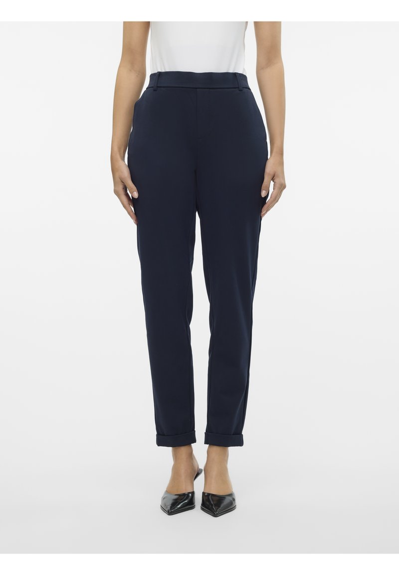 Femme portant un pantalon bleu marine fuselé avec des revers et des chaussures noires à bout pointu sans lacets, debout devant un fond blanc.