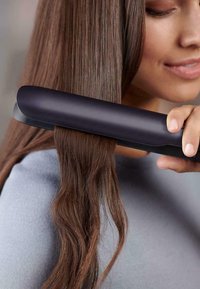 Glätteisen mit einem eleganten schwarzen Gehäuse, das sanft über lange, glänzende braune Haare gleitet und weiche Wellen kreiert. Glatte Metallplatten und abgerundete Kanten.