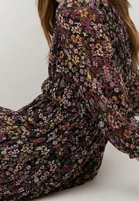 Květinové šaty z tmavé látky s růžovými, žlutými a bílými květinami. Dlouhé rukávy, plisé design a lehká textura.