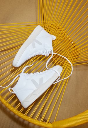 Baskets montantes blanches avec logo gris « V » et lacets, placées sur une chaise à cordes jaune sur un fond neutre.