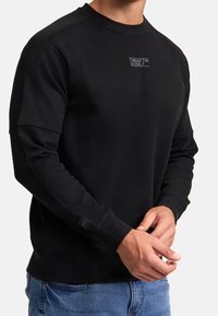 Mand iført en sort langærmet sweatshirt med teksten "WEAR THE CODE" på brystet og blå jeans, hænderne foldet foran.